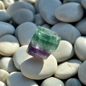 A rough, cylindrical fluorite crystal with green and purple bands rests on smooth white pebbles, bathed in soft sunlight, creating a serene scene.